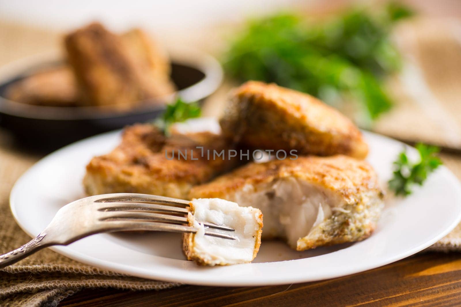 A piece of hake fish fried in a crispy crust, in a plate on a wooden table.