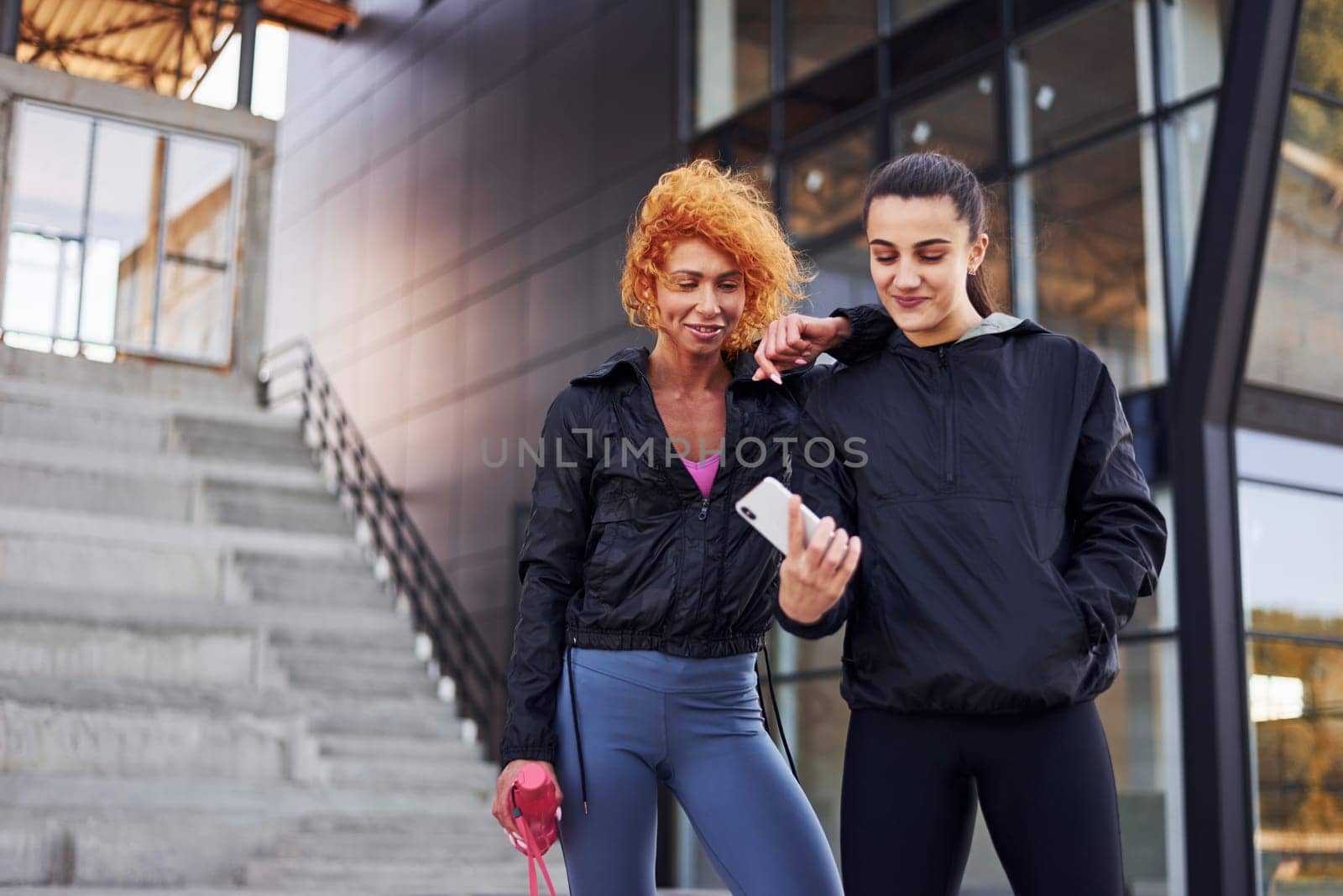 Holds phone in hands. Two female friends with sportive bodies have fitness day outdoors.
