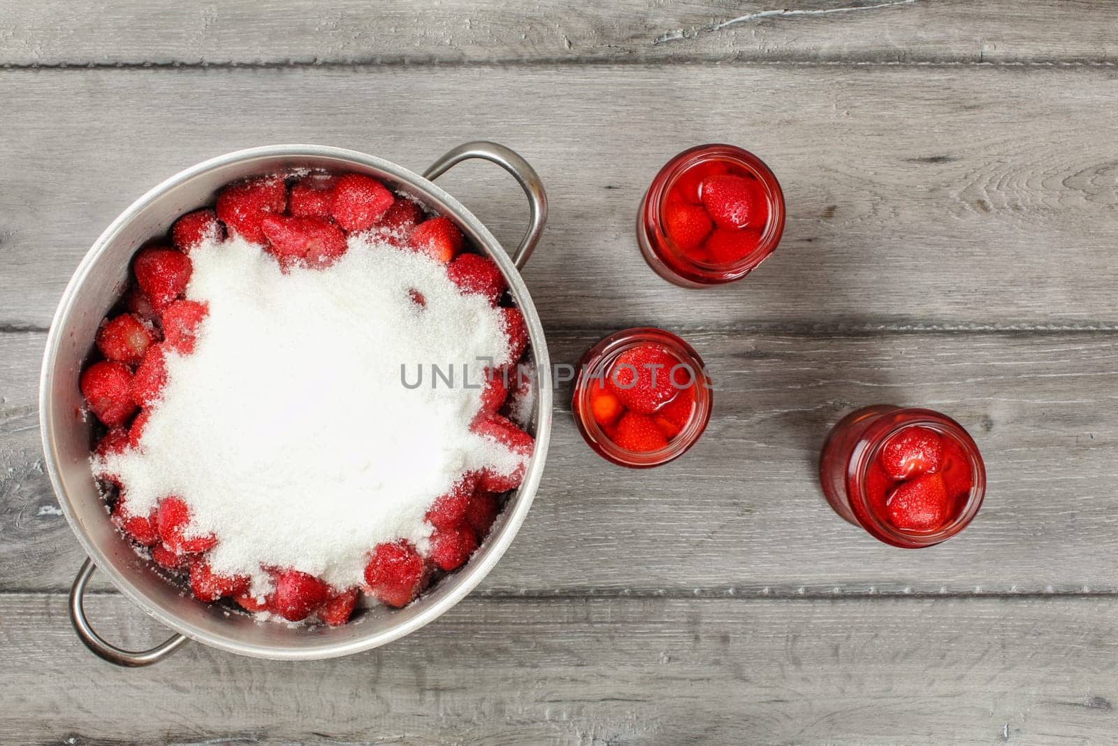 Tabletop view, large steel pot full of strawberries covered with crystal sugar, three bottles of strawberry in syrup next to it on gray wood desk. Homemade compote preparation.
