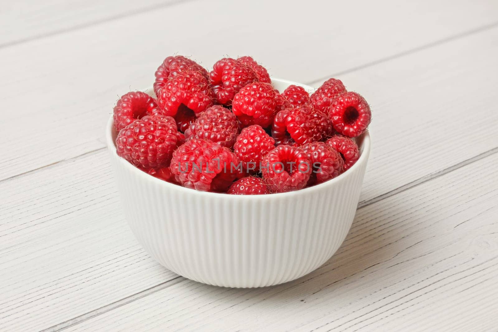 Small white ceramic bowl full of freshly picked raspberries, light boards desk under.
