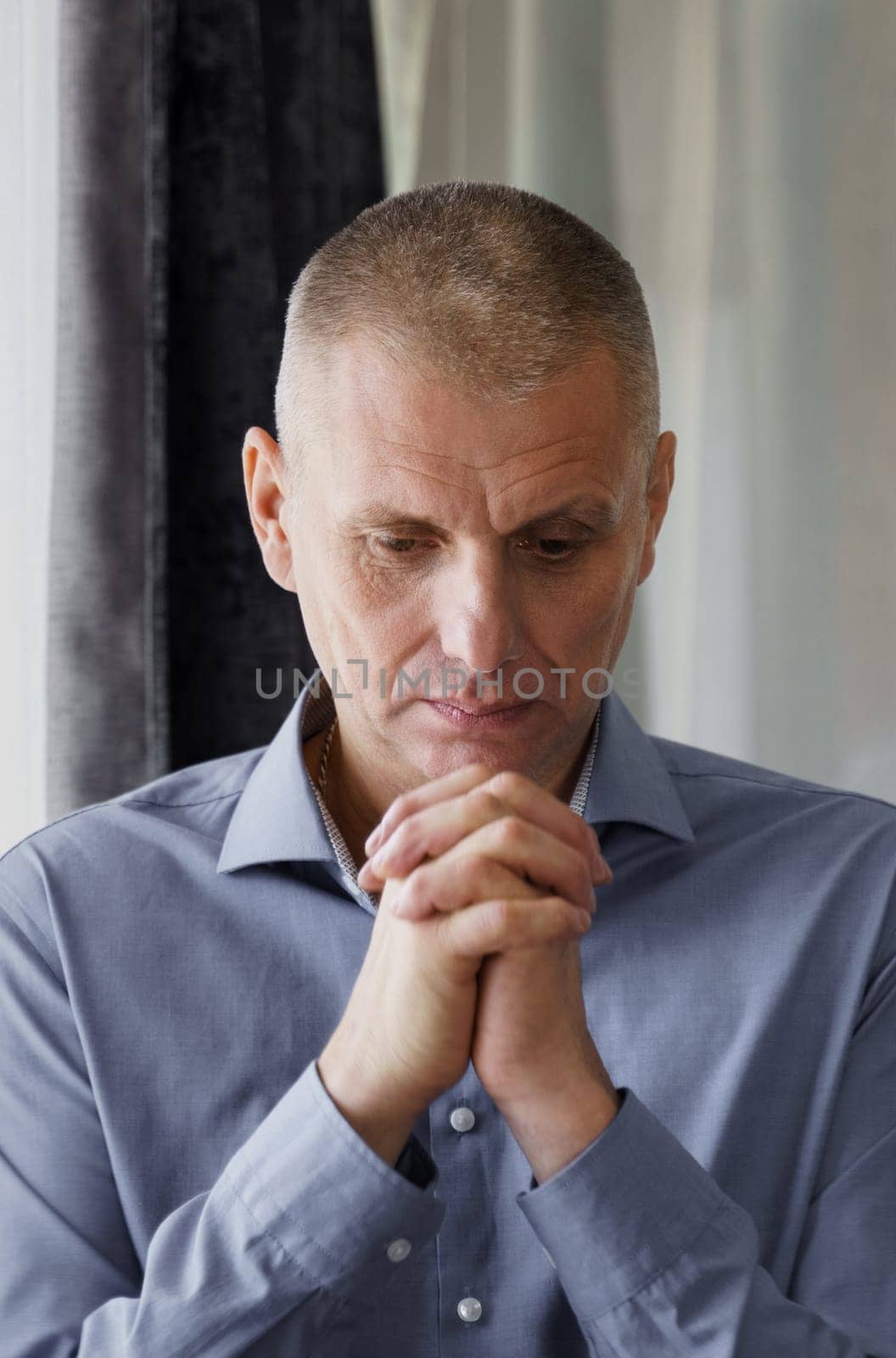 Portrait of a man who stands at the window clenched his hands in front of him, concentrated, prays or thinks. Vertical frame.