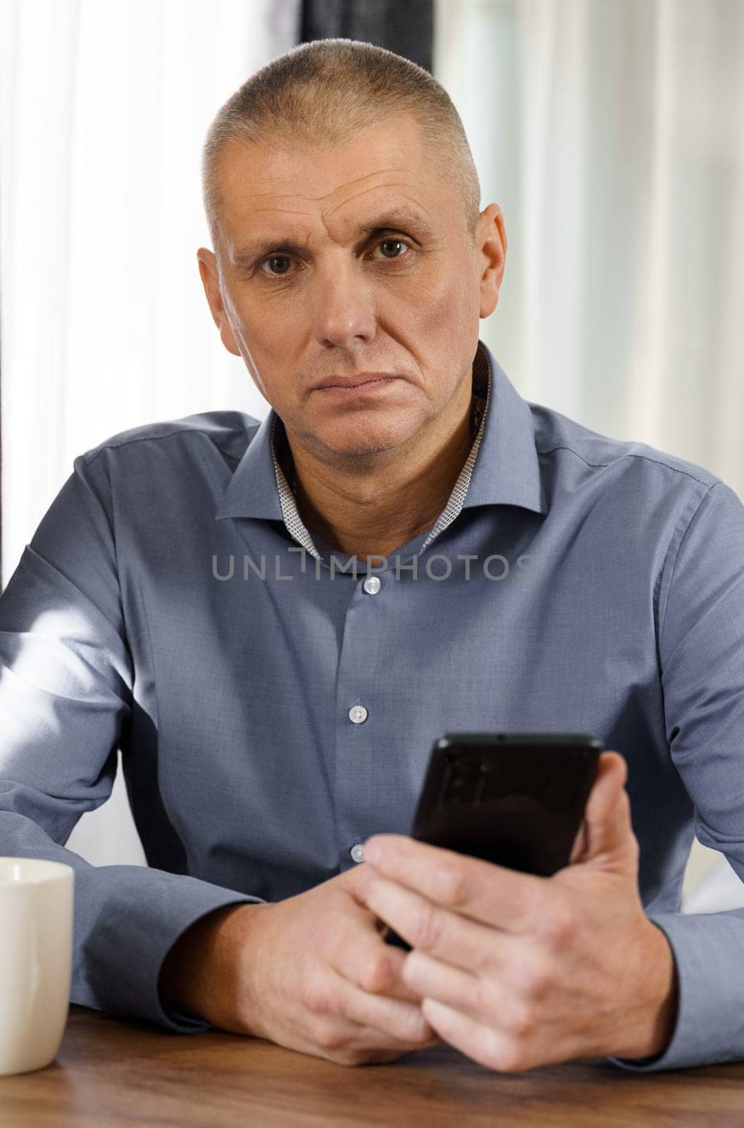 Portrait of a male businessman who sits at a table, holds a phone in his hands, looks into the camera. Vertical frame.