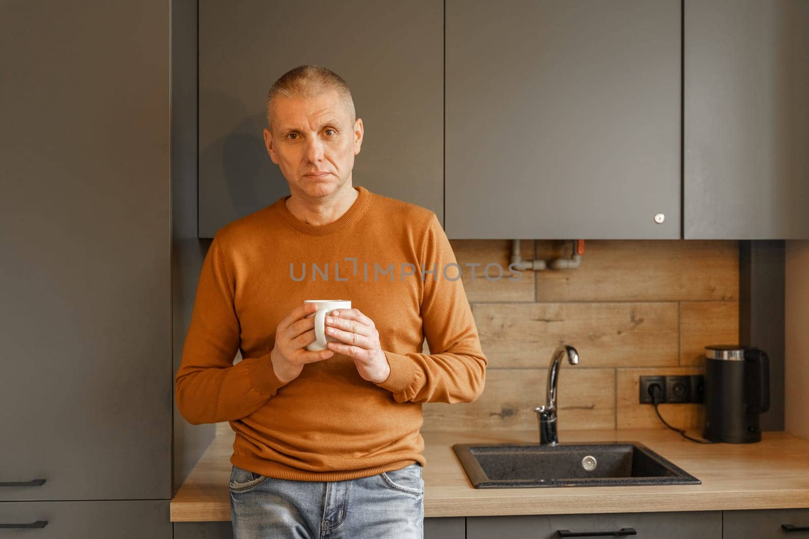 Portrait of a mature man in an orange jumper in the kitchen with a mug of warm tea.