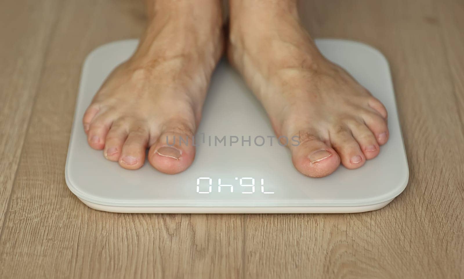 Man weighing himself - male bare feet stepping on white digital floor scales at home: close up view. Measuring weight, control, wellness and diet concept.