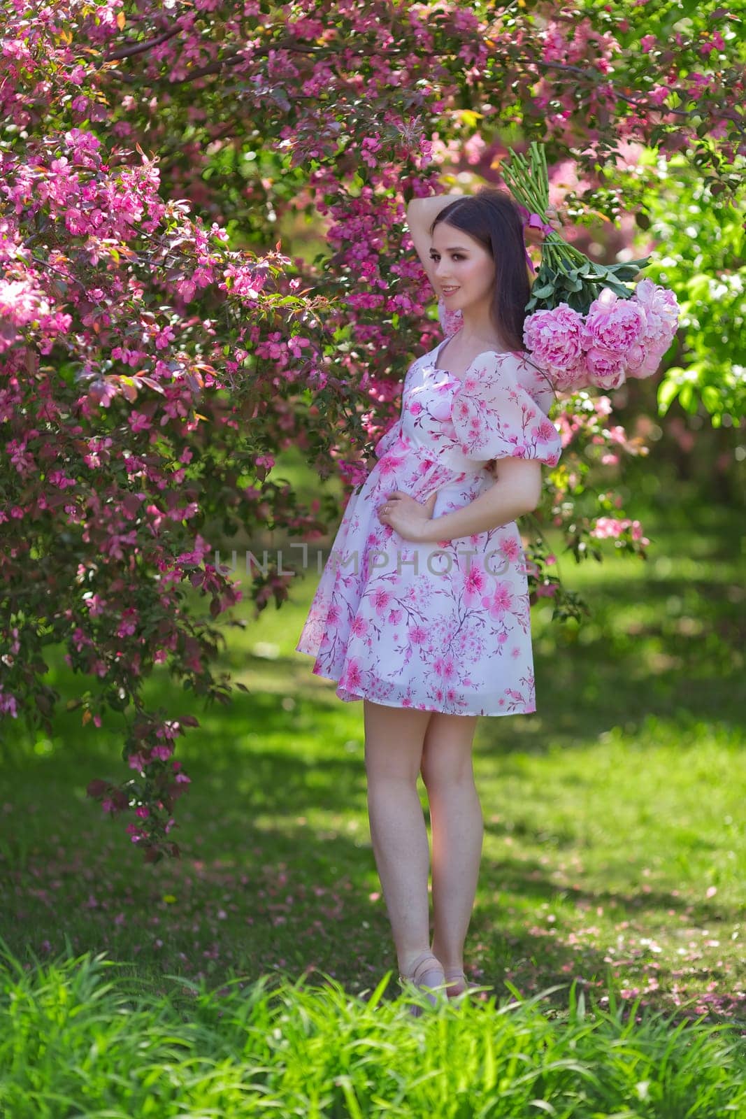 A beautiful happy girl with long hair, a brunette, in a light pink dress, holding a bouquet of large pink peonies behind her back, standing near pink flowering apple trees, in the garden on a sunny day in spring. Vertical