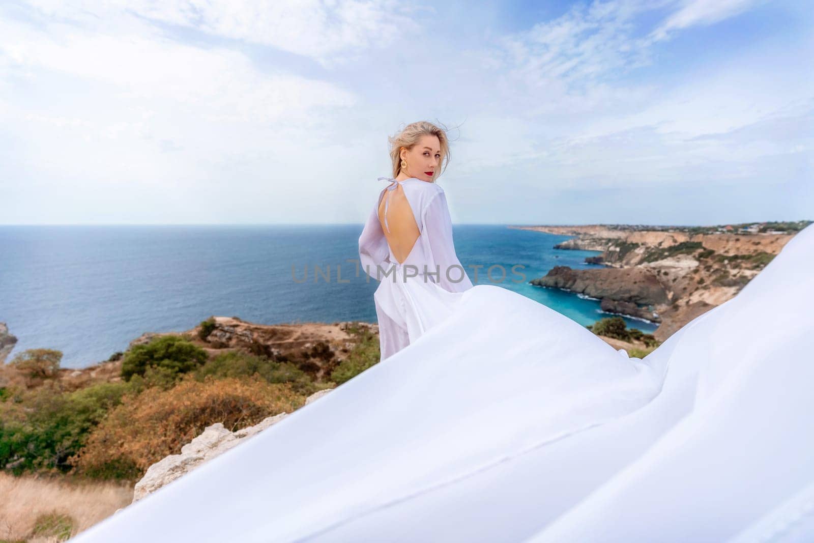 Blonde with long hair on a sunny seashore in a white flowing dress, rear view, silk fabric waving in the wind. Against the backdrop of the blue sky and mountains on the seashore