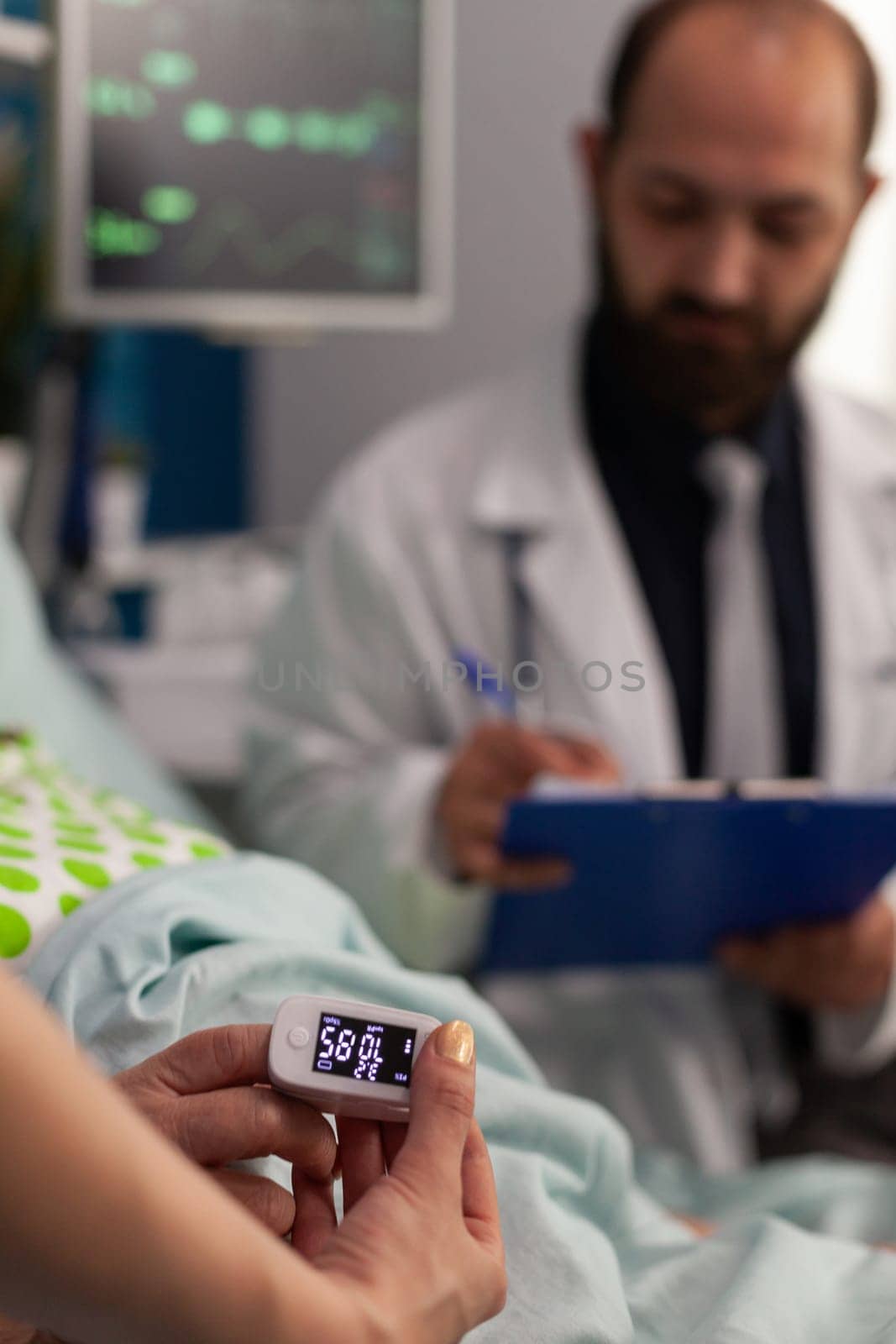 Nurse measuring blood oxygen level with oximeter on elderly patient lying in hospital bed. Male doctor reviewing results of medical examinations of retired woman, specialized care.