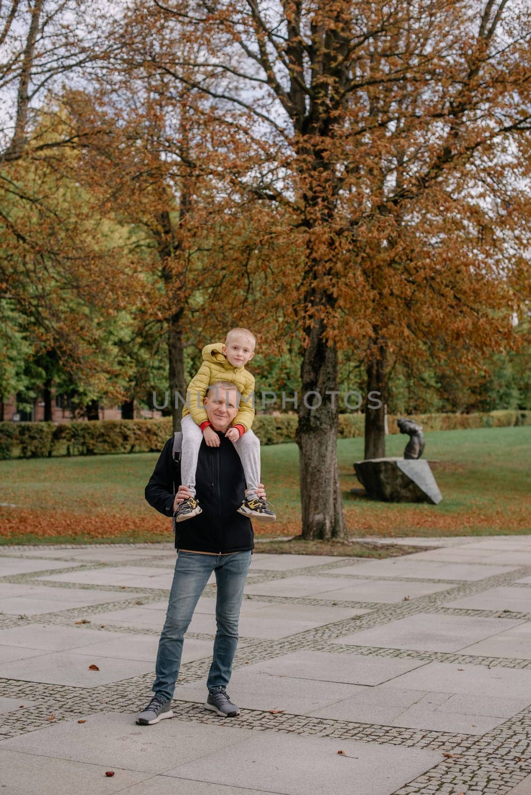 Dad holds on his son's shoulders. Beautiful family is spending time together outside. Dad and his little son are having fun on a roof terrace with view on a city. Sitting on father's shoulders and smiling. by Andrii_Ko