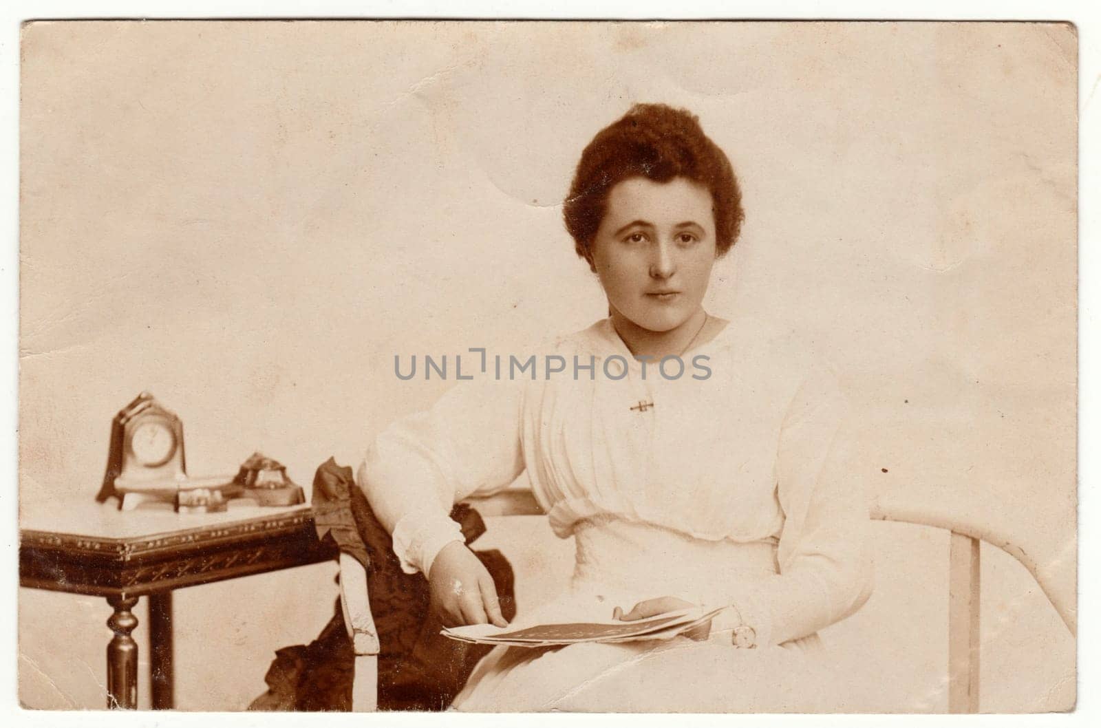 POZNAÅ  (POSEN), POLAND - CIRCA 1920s: Vintage photo shows a young woman. She wears a white dress. Historical table and an old world mantel clock are on the background. Retro black and white studio photography. Circa 1920s.