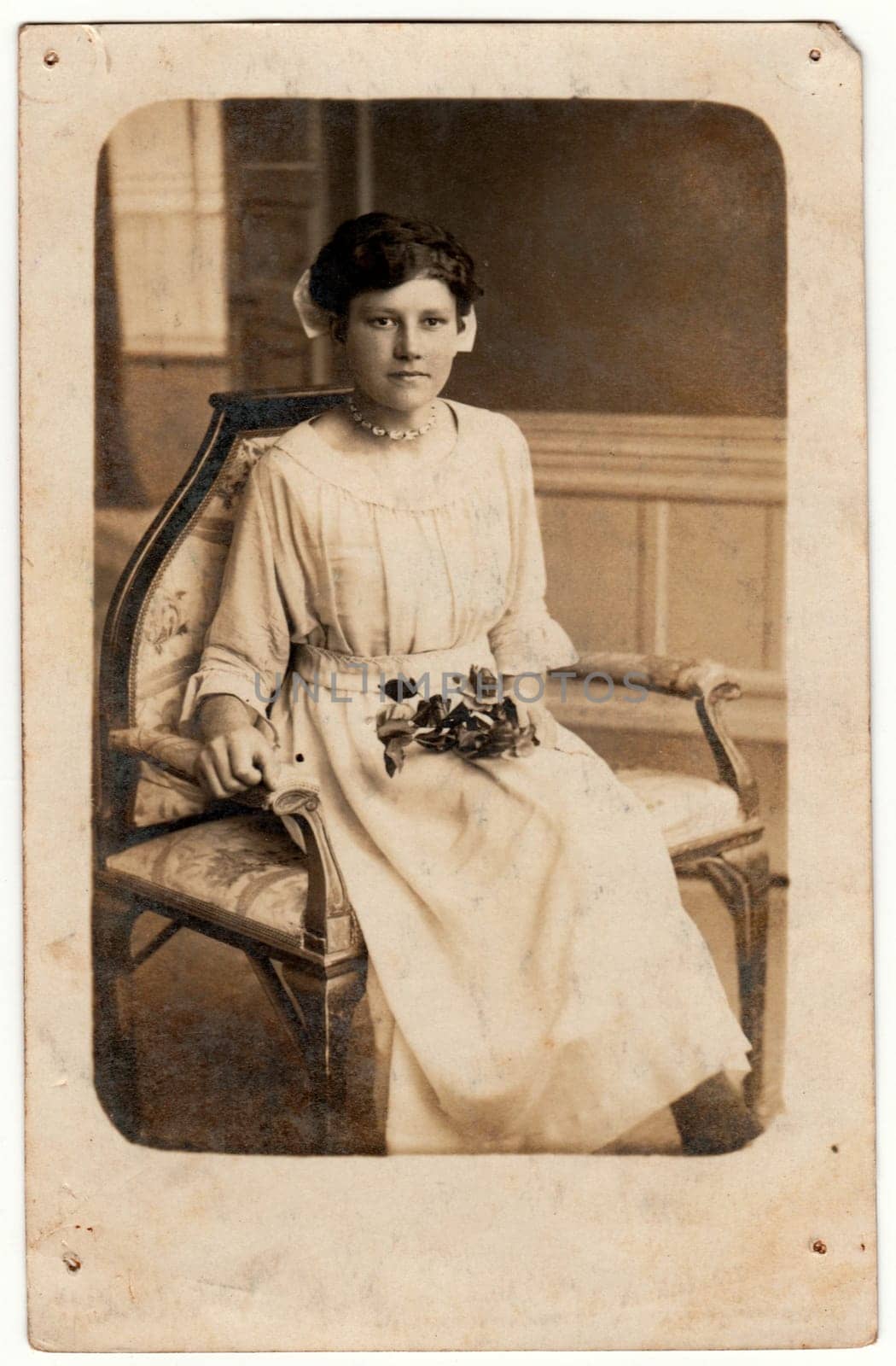 Vintage photo shows a young woman. She wears a white dress. She sits on the historical armchair and holds roses. Retro black and white studio photography. by roman_nerud