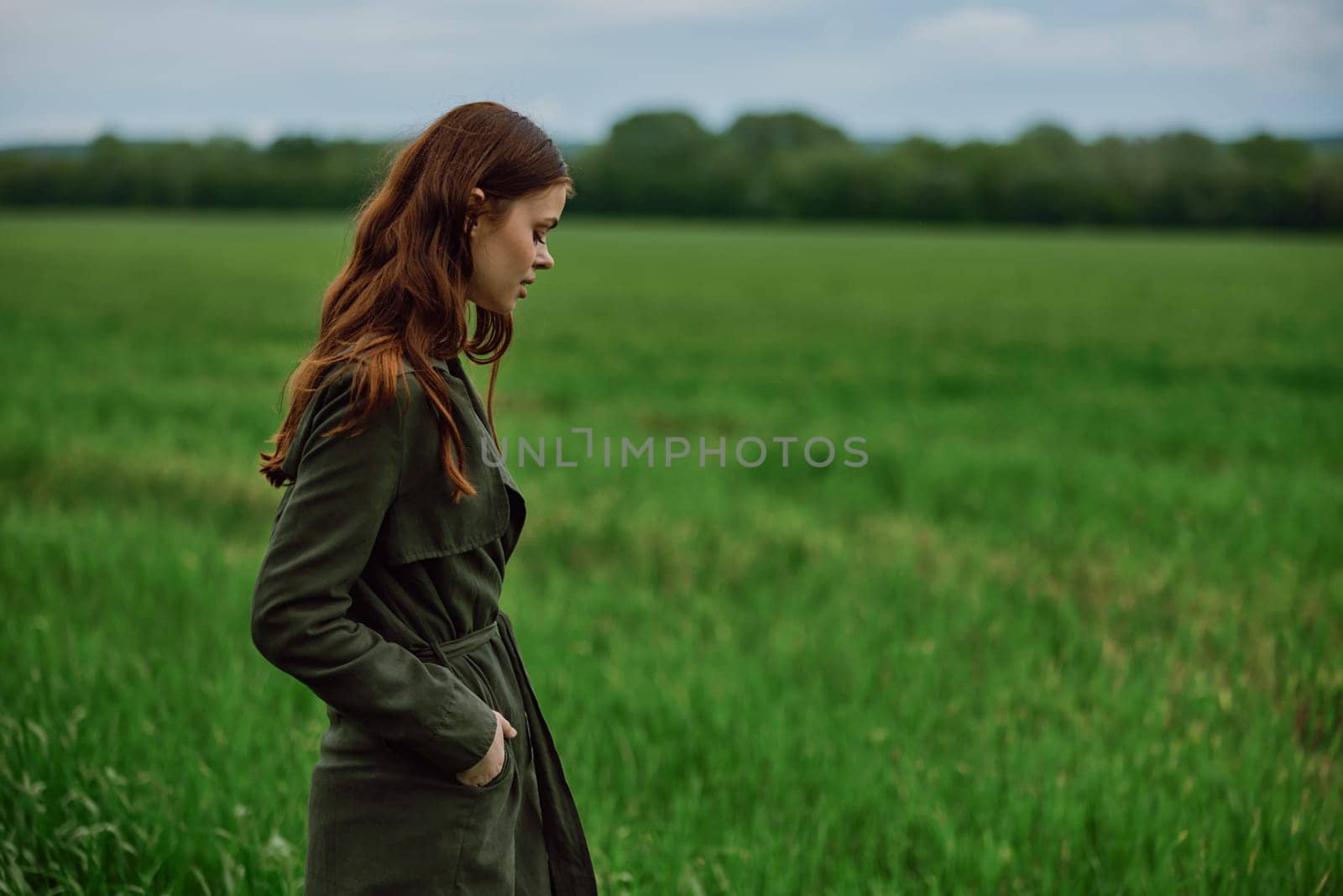 a beautiful, red-haired girl in a raincoat stands in a field in the spring in rainy weather by Vichizh