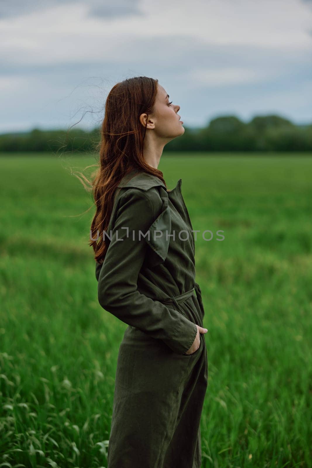 a beautiful red-haired woman stands in a field in a long dark raincoat, holding her hands in her pockets by Vichizh