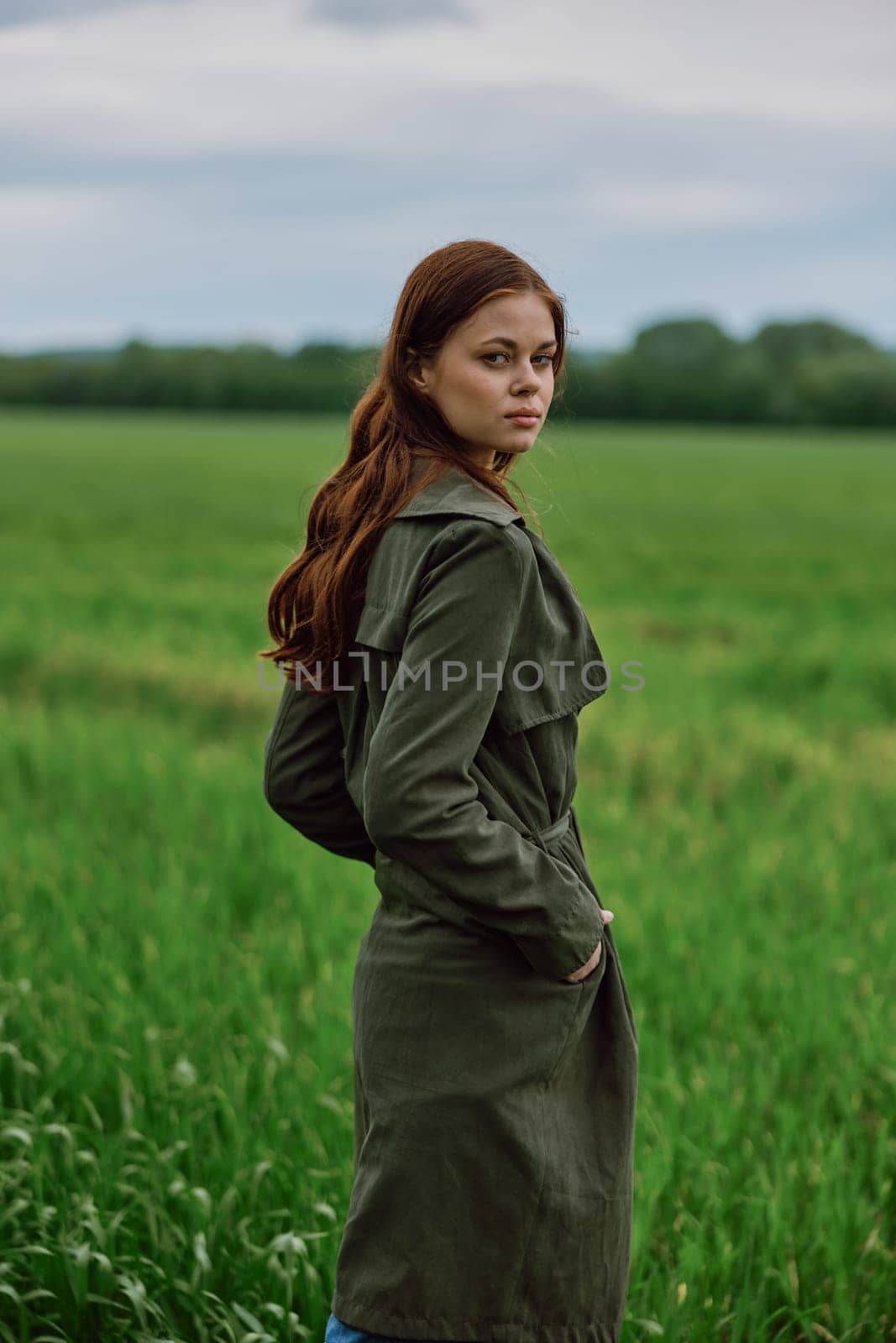 a beautiful red-haired woman stands in a field in a long dark raincoat, holding her hands in her pockets. High quality photo