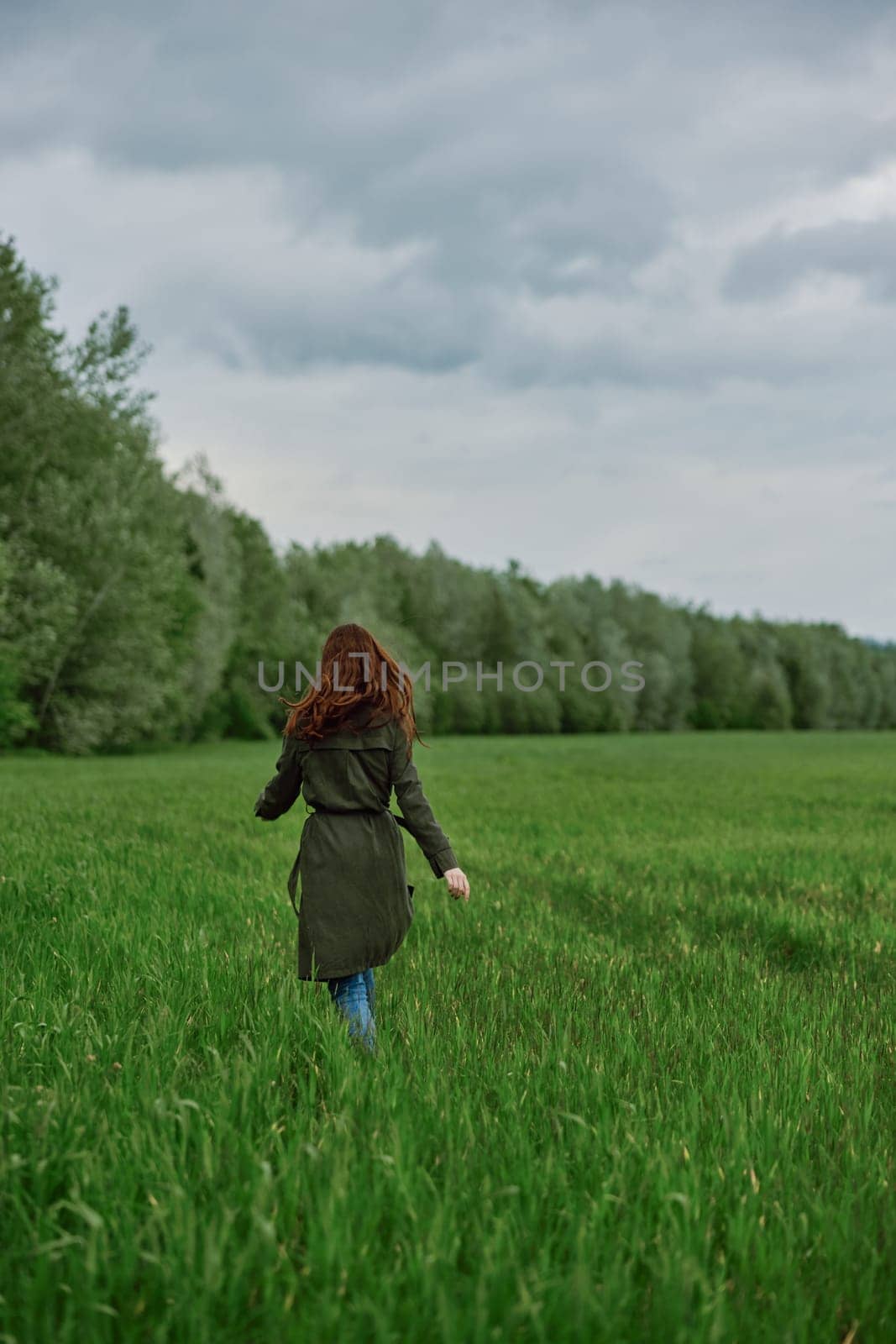 a woman in a long raincoat runs across a field in tall green grass in cloudy weather in spring by Vichizh