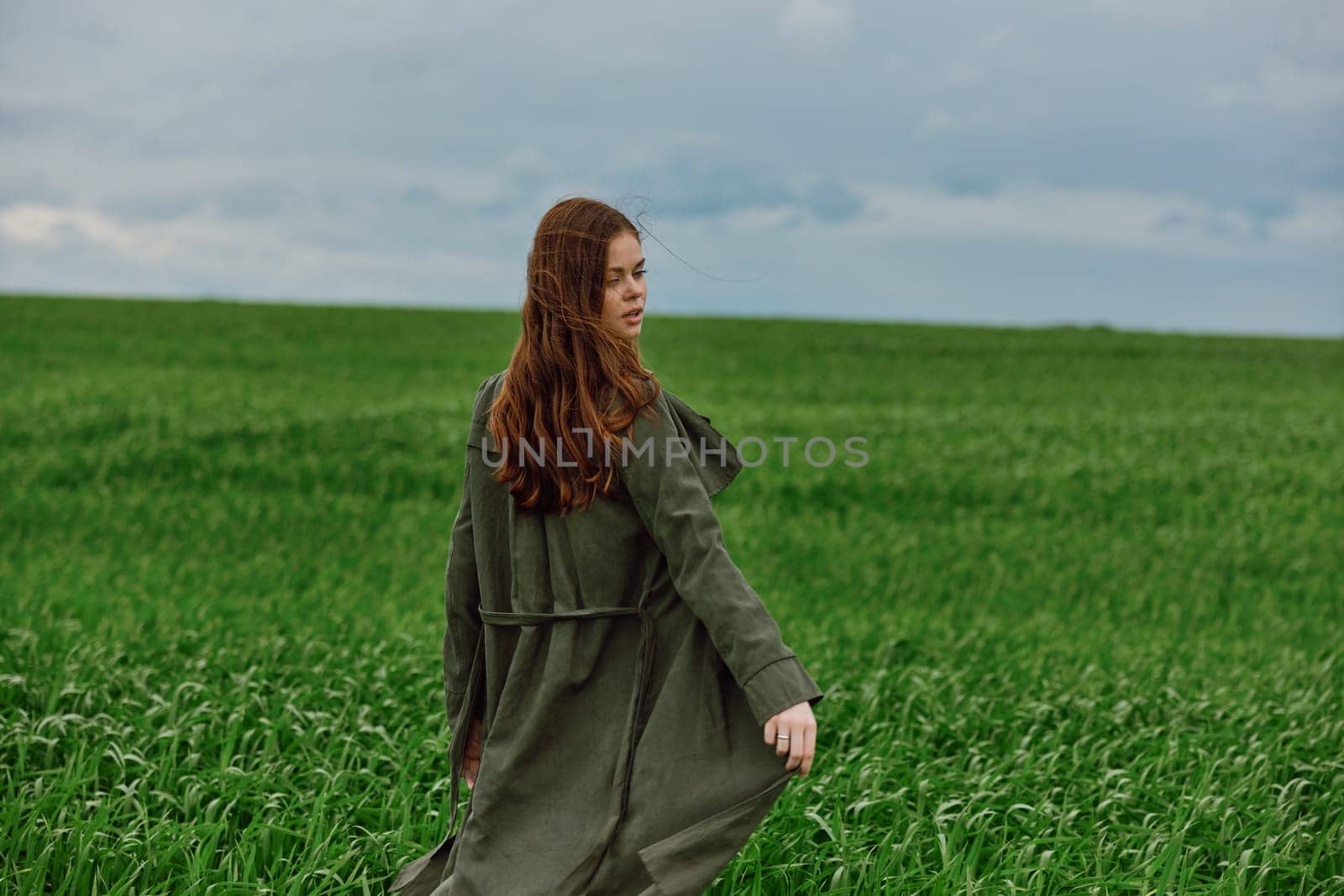 a woman in a dark coat stands in a green field in the wind close to the camera. Strong wind, cloudy weather. High quality photo