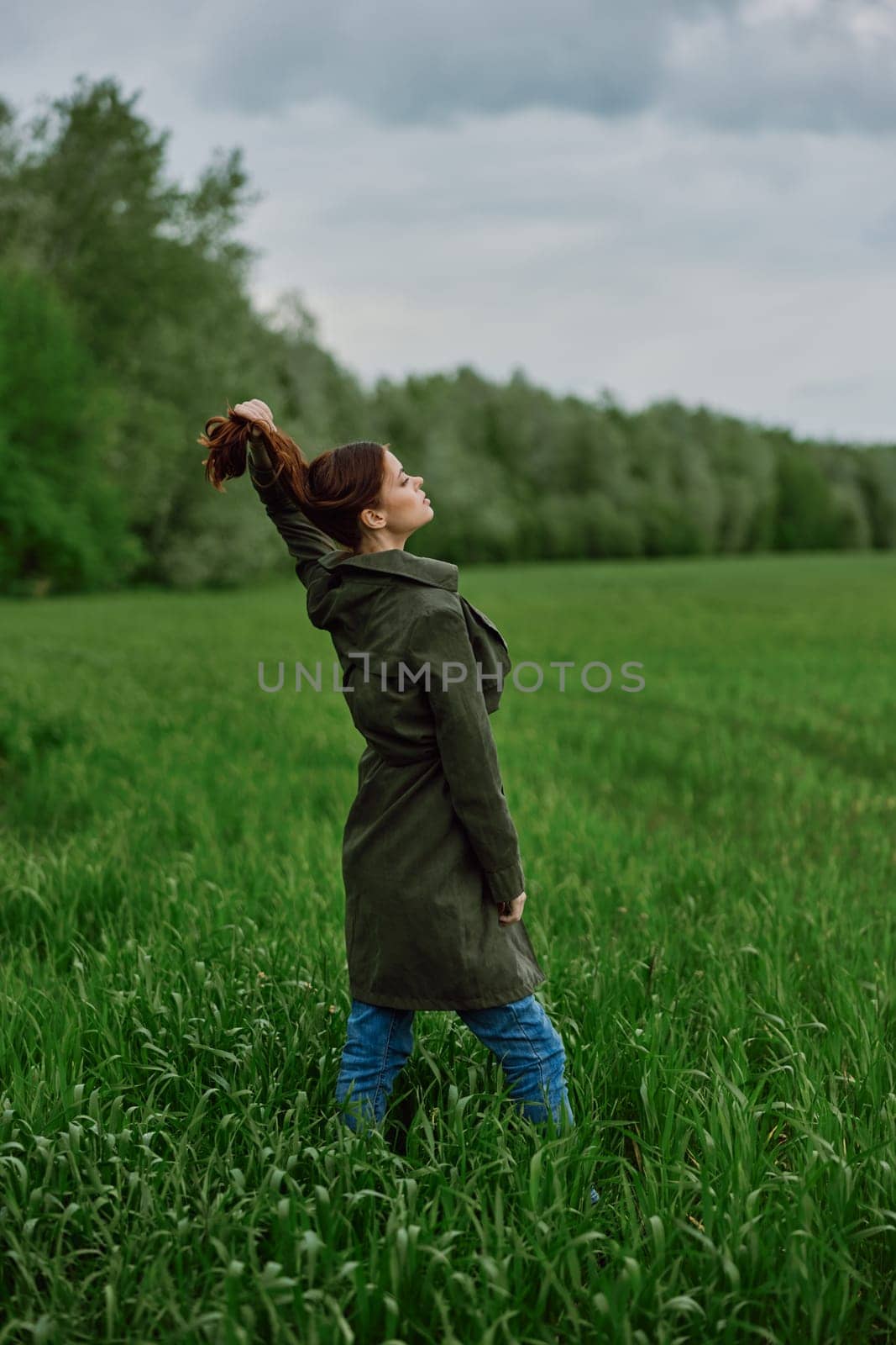 a beautiful woman in a dark coat stands in a green field in the spring in rainy weather and straightens her hair by Vichizh