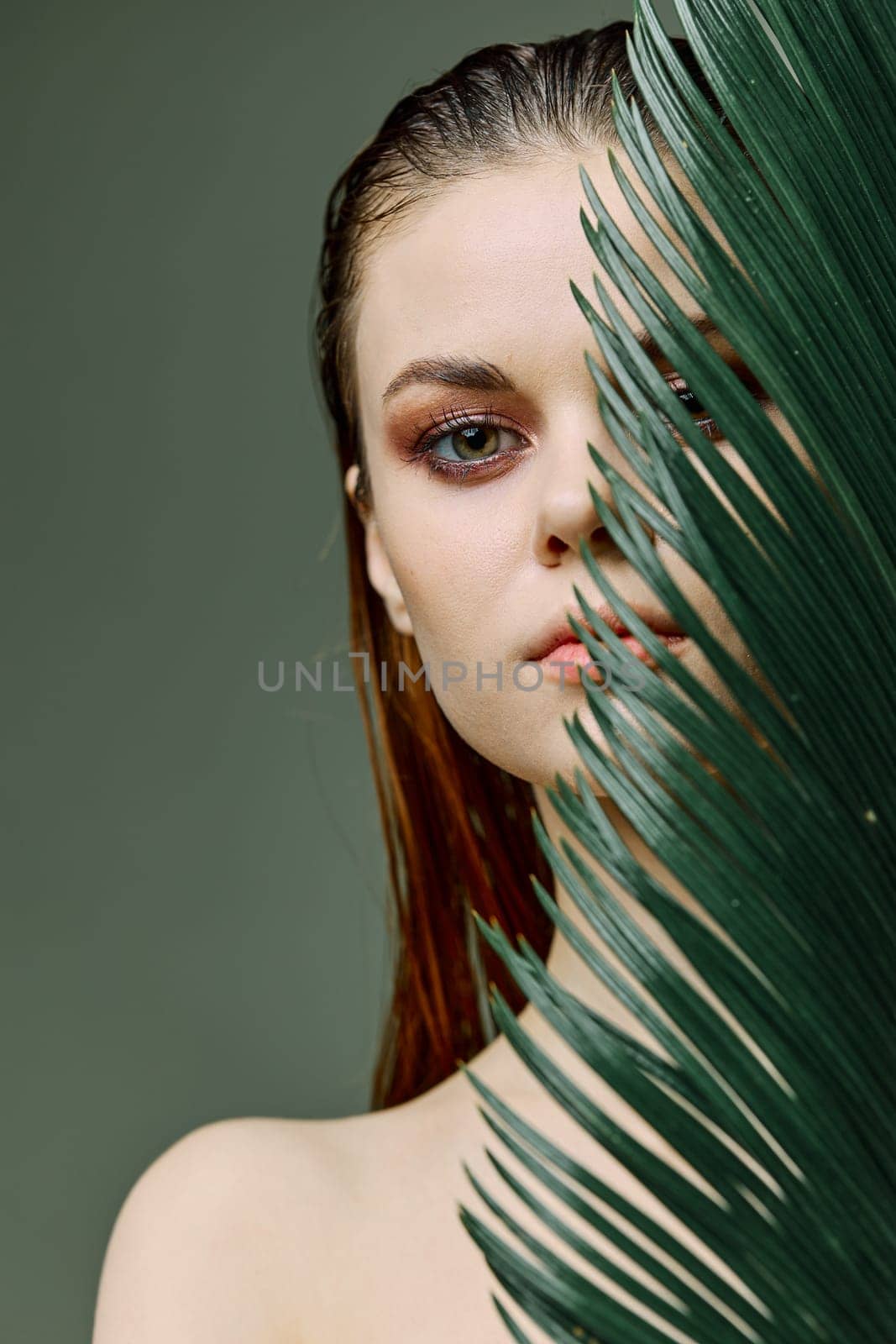 a sophisticated, elegant woman with evening makeup, stands with a green palm leaf, covering part of her face with it. Vertical photo without retouching. High quality photo