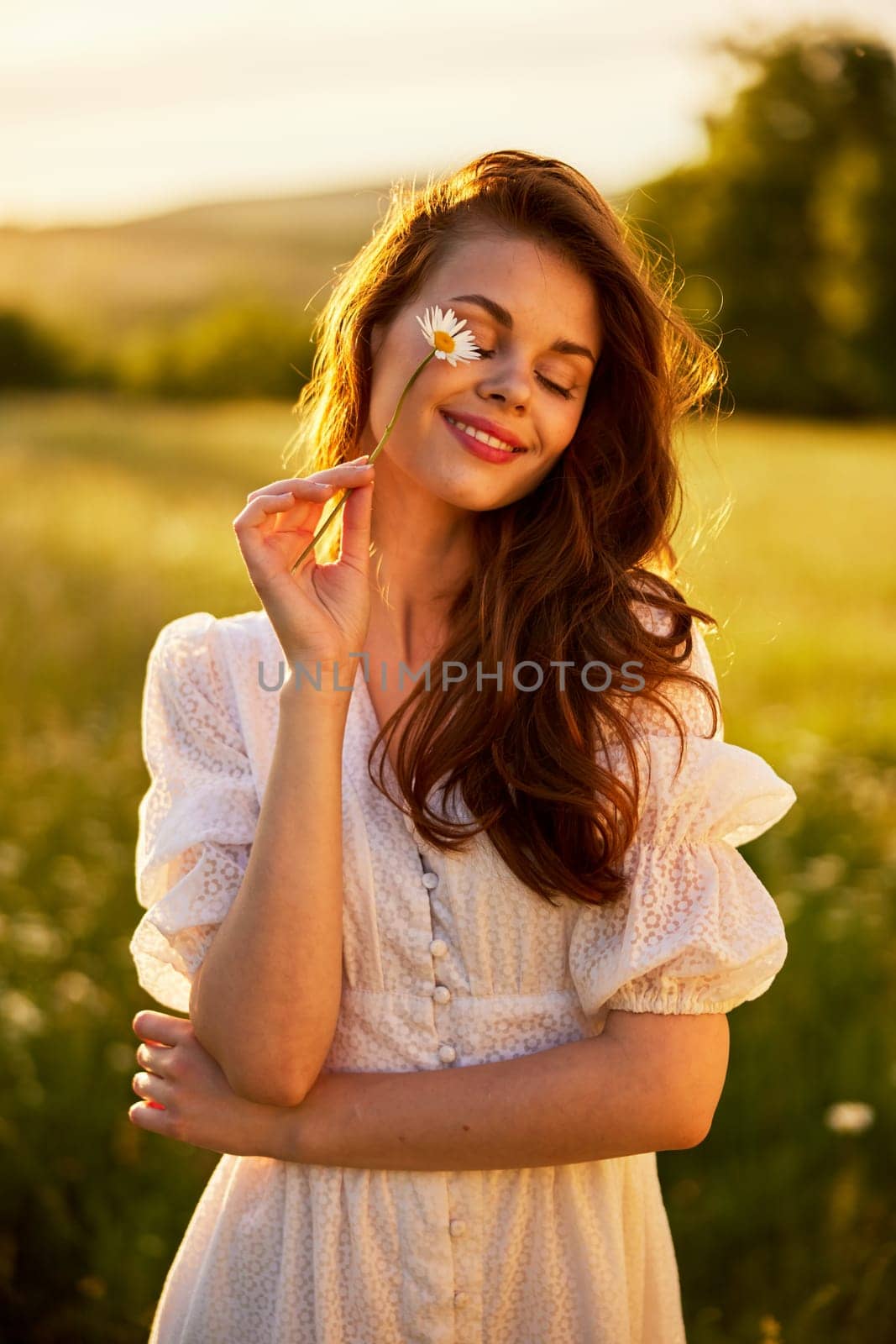 close portrait of a beautiful, laughing woman in a light dress holding a chamomile near her face during sunset, lit from the back. High quality photo