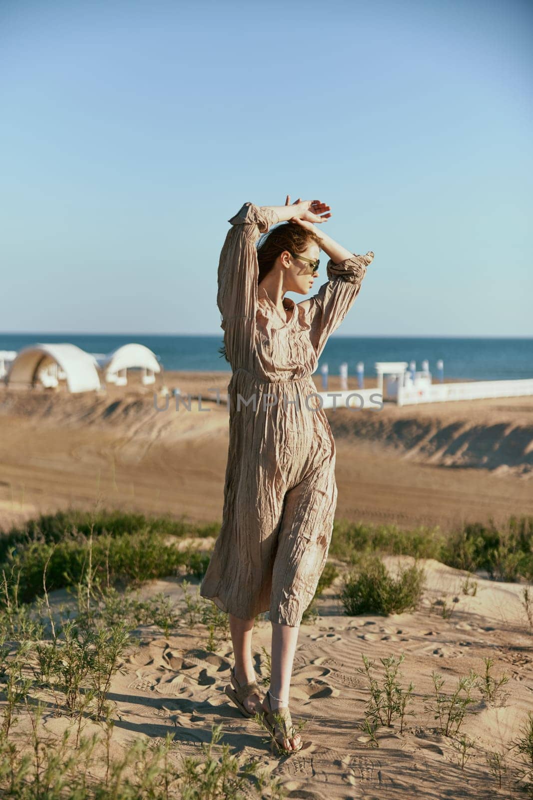 a slender, sophisticated woman in a beige dress stands on the sand in windy weather with her hands raised to her head. High quality photo