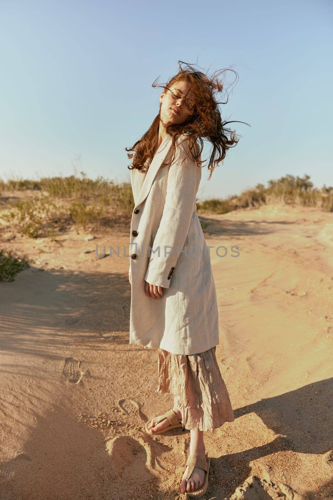 a woman poses standing on the sand in light summer clothes against the blue sky and the wind blows her hair on her face. High quality photo