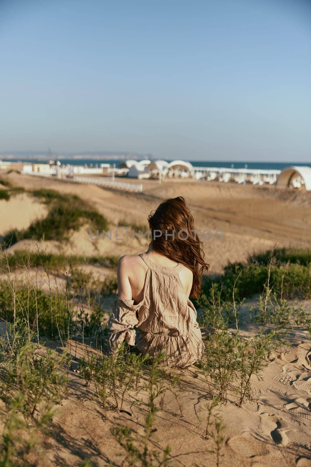 red-haired woman posing sitting with her back to the camera in a beige dress on the sea coast during sunset. High quality photo