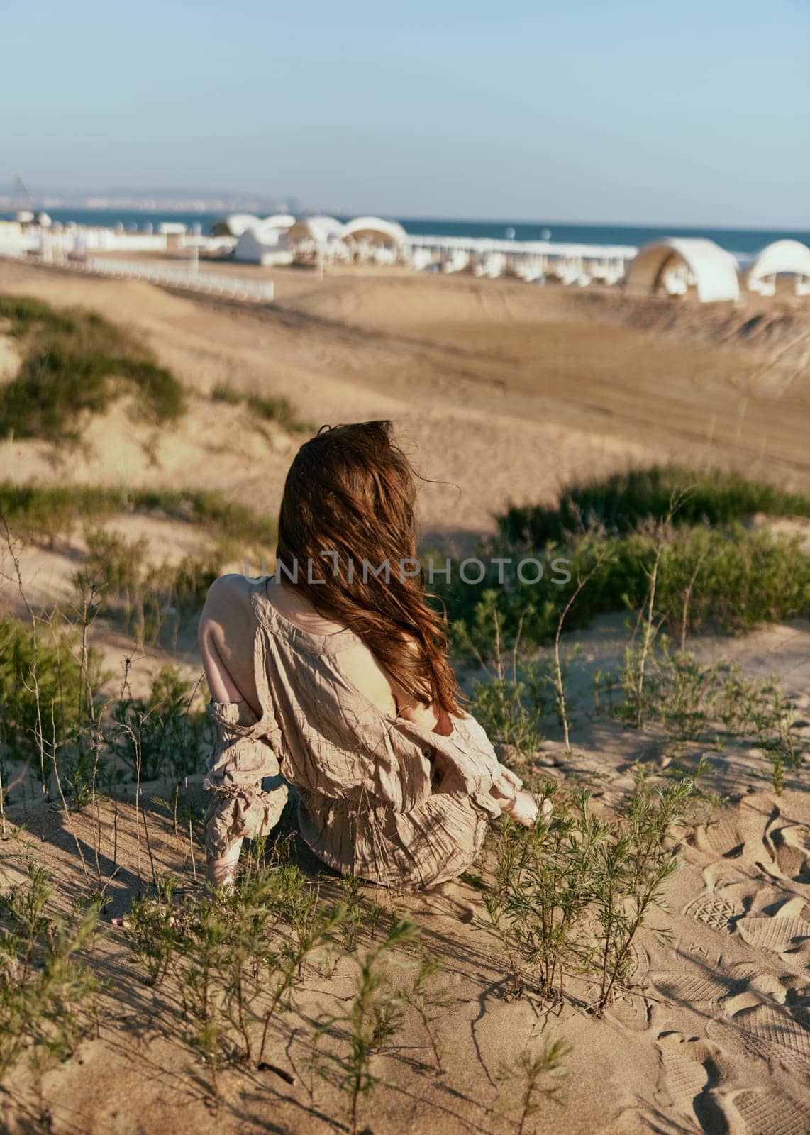 red-haired woman posing sitting with her back to the camera in a beige dress on the sea coast during sunset. High quality photo