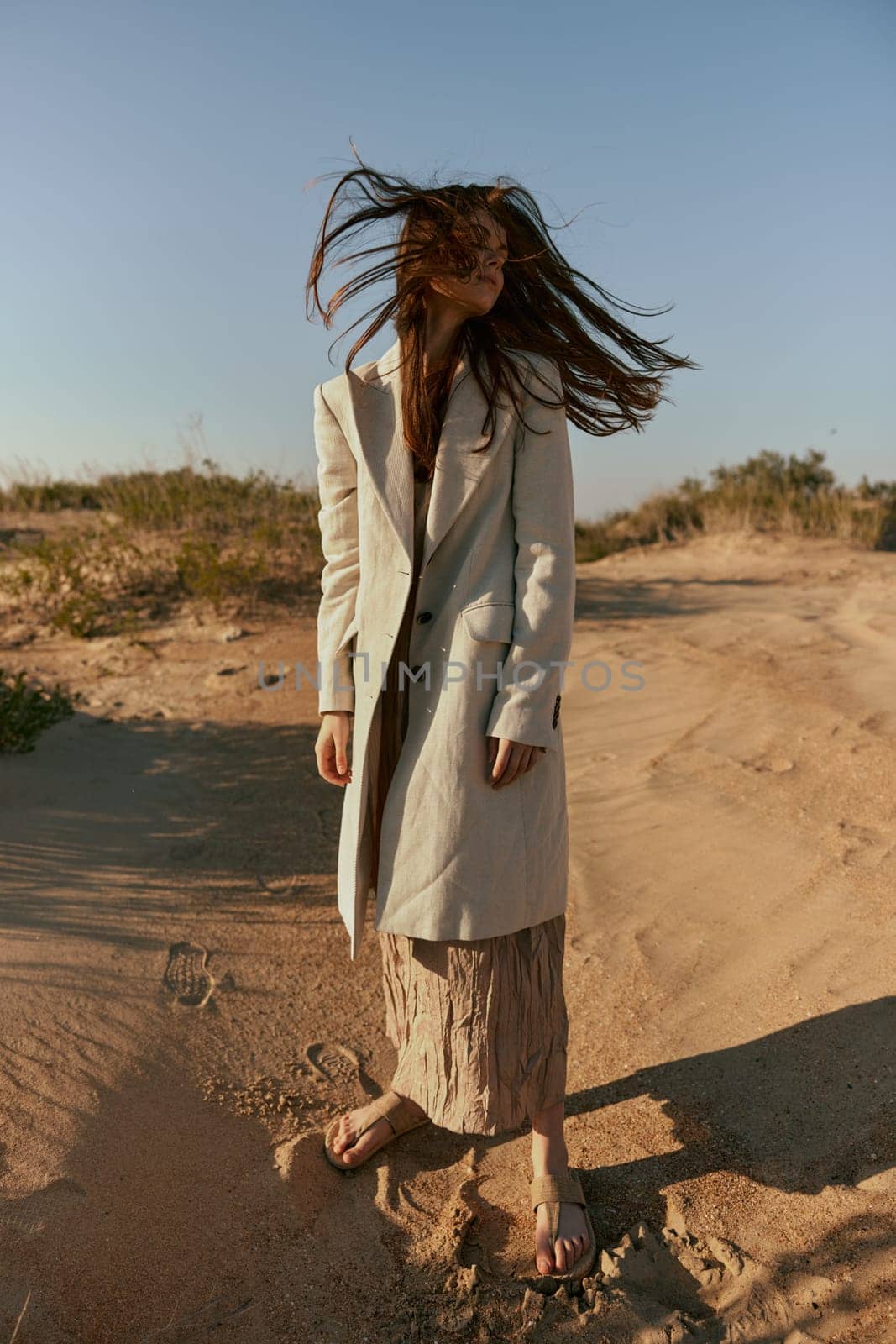 a woman poses standing on the sand in light summer clothes against the blue sky and the wind blows her hair on her face. High quality photo