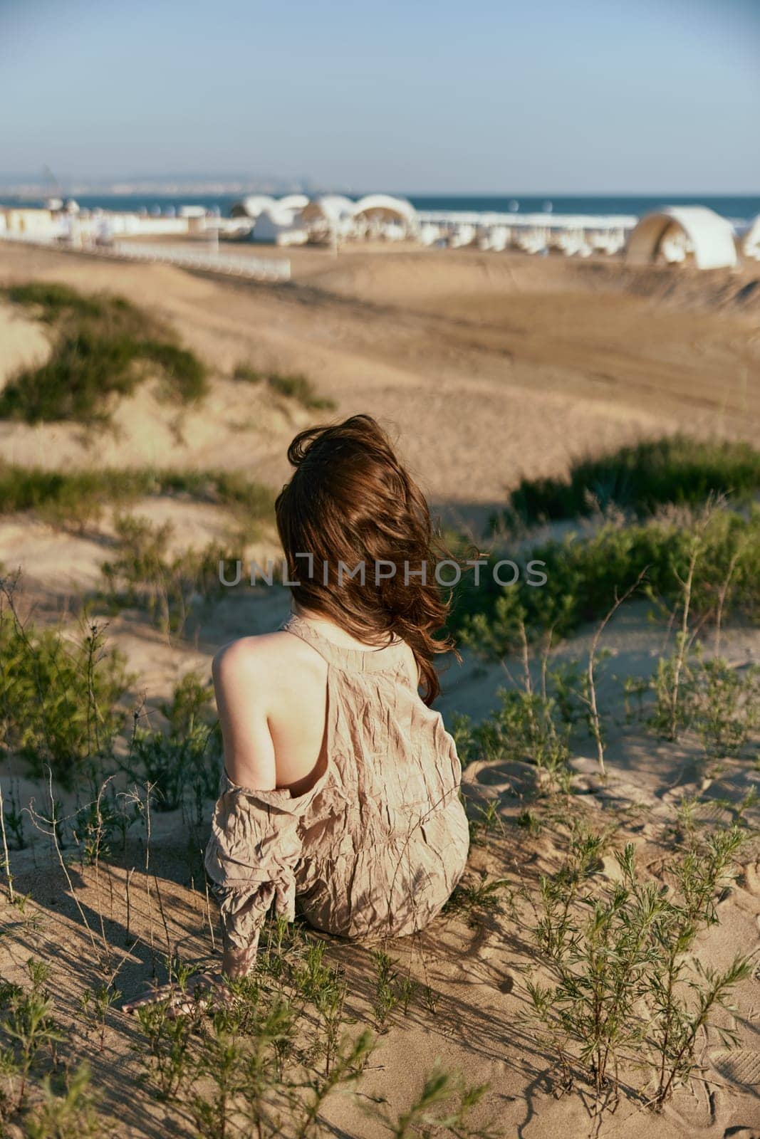 red-haired woman posing sitting with her back to the camera in a beige dress on the sea coast during sunset. High quality photo