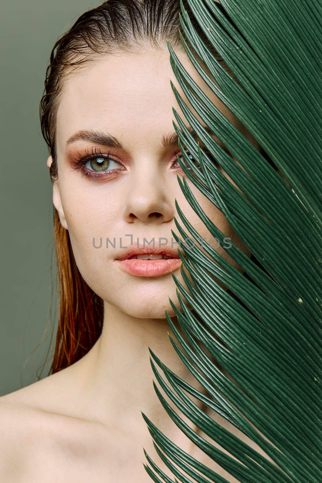 a sophisticated, elegant woman with evening makeup, stands with a green palm leaf, covering part of her face with it. Close vertical portrait, photo without retouching by Vichizh