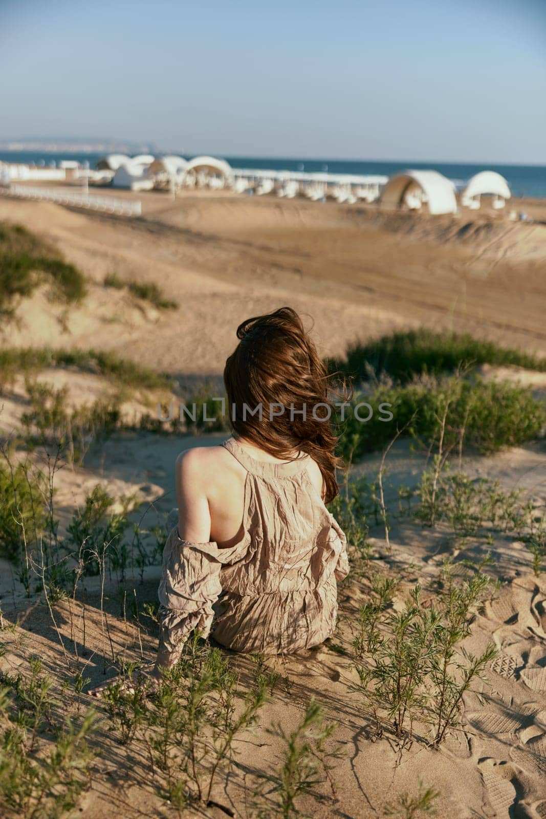 red-haired woman posing sitting with her back to the camera in a beige dress on the sea coast during sunset. High quality photo