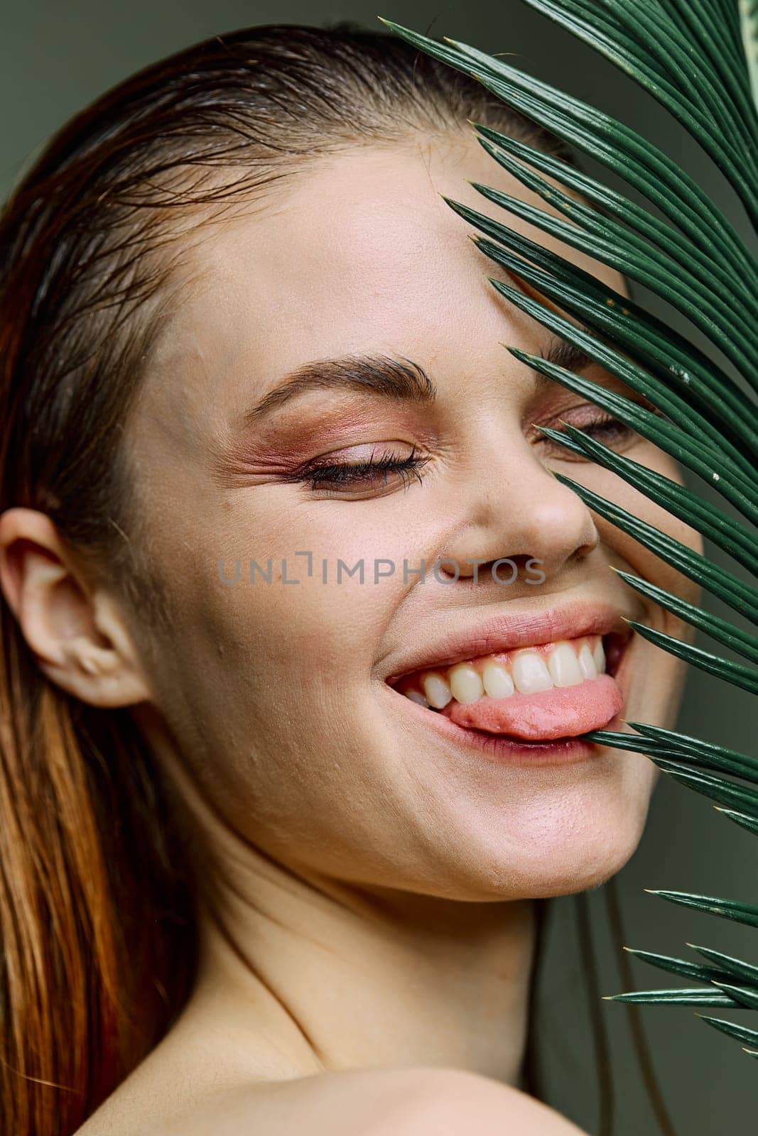 beauty portrait of a beautiful, elegant woman standing holding a palm leaf near her face, amusingly showing her tongue. Vertical photo without retouching of problem skin. High quality photo