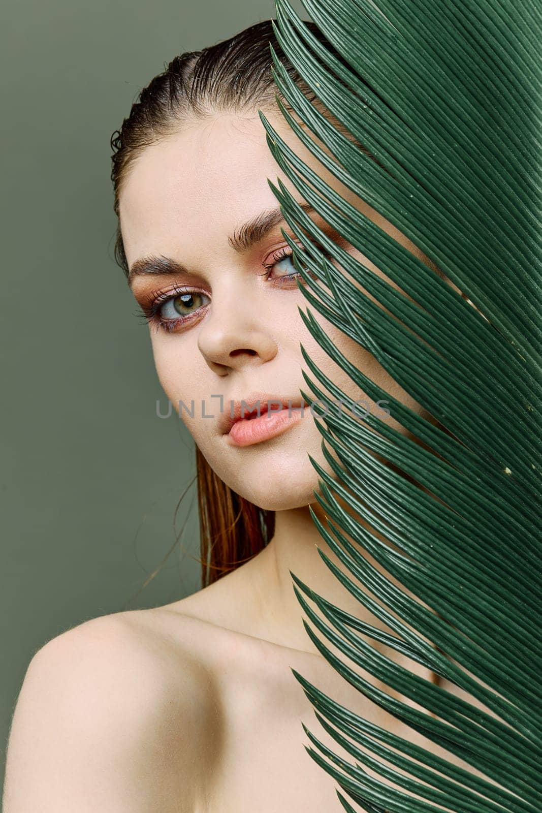a sophisticated, elegant woman with evening makeup, stands with a green palm leaf, covering part of her face with it. Vertical photo without retouching by Vichizh