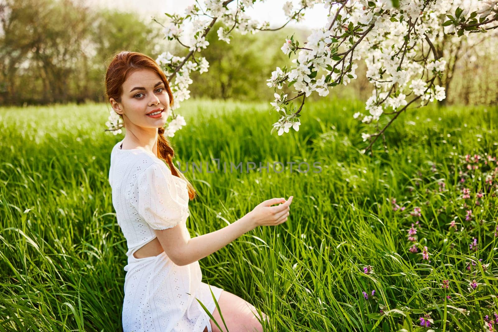 a lovely woman in a light short dress sits near a flowering tree and smiles while looking at the camera by Vichizh