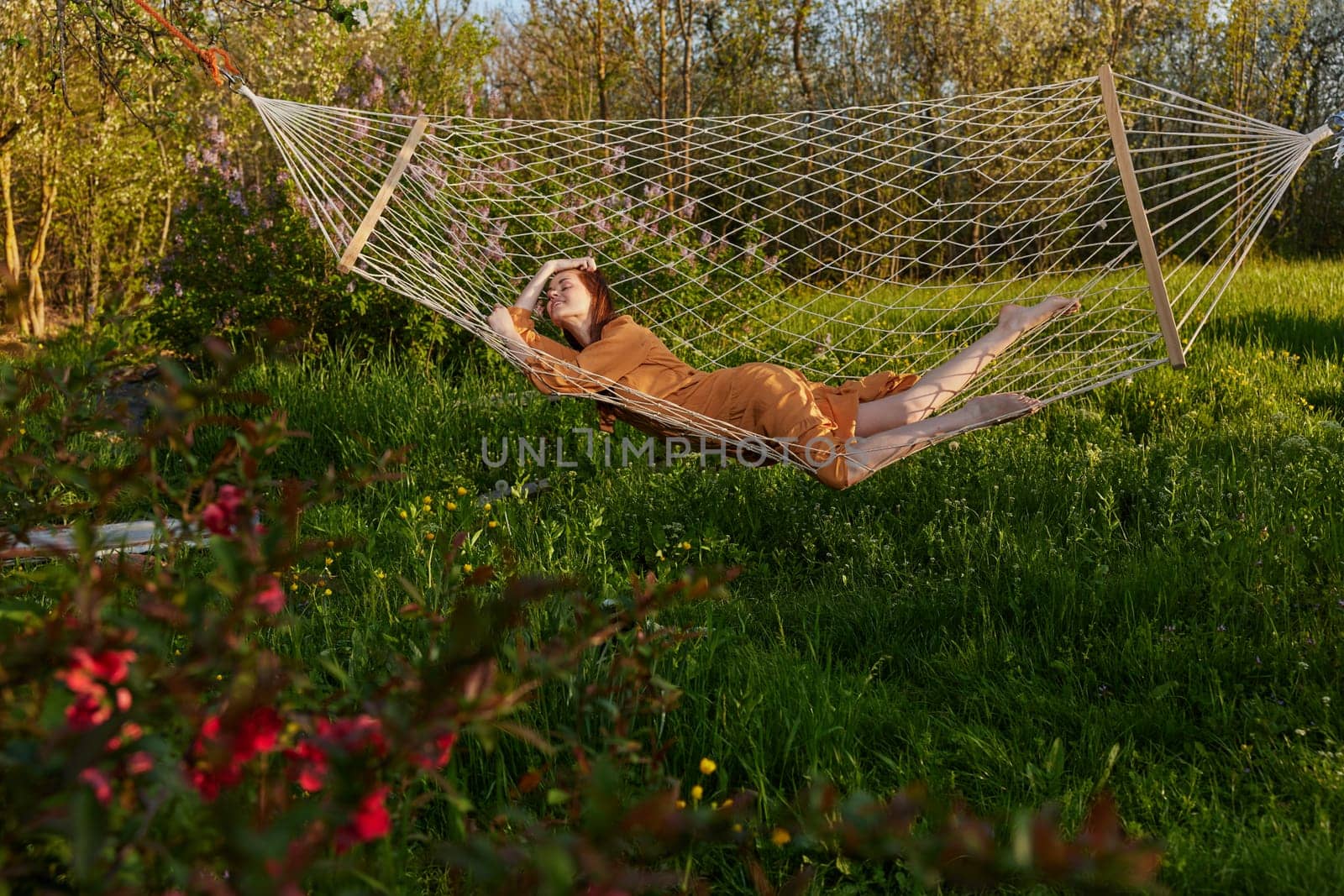 a happy woman in a long orange dress is relaxing in nature lying in a mesh hammock enjoying summer and vacation in the country surrounded by green foliage. High quality photo