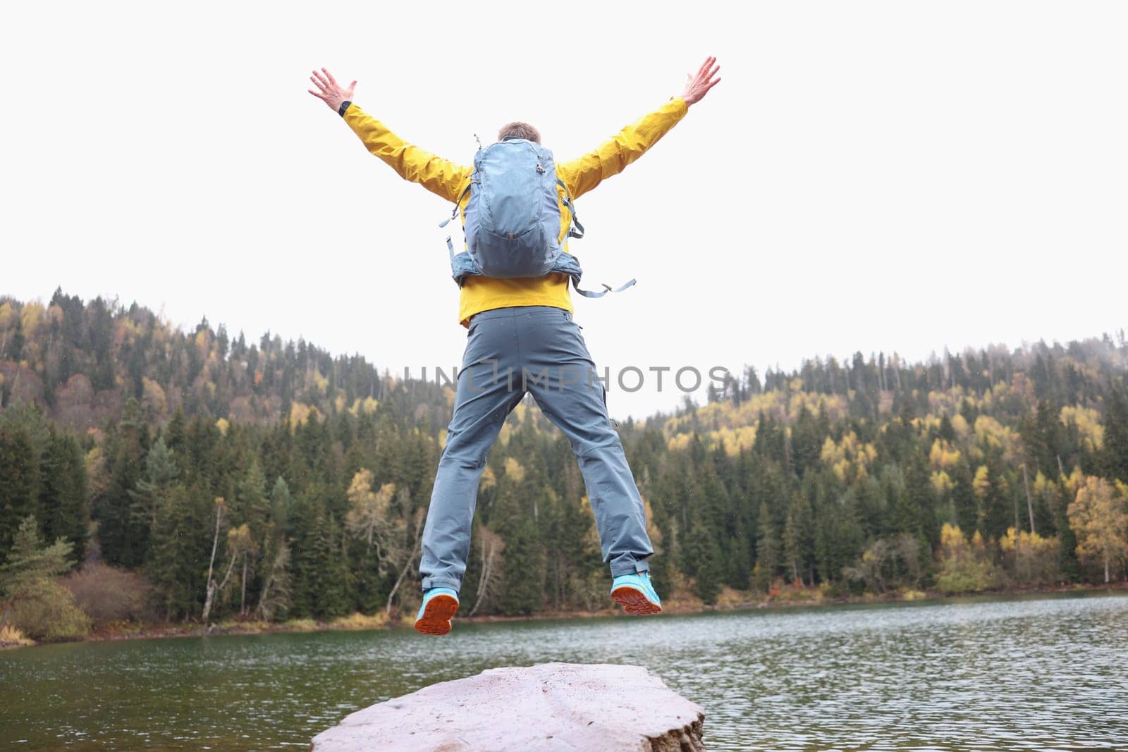 Happy male traveler with backpack jumping near forest lake while reaching goal back view closeup. Freedom and fulfillment of desires concept