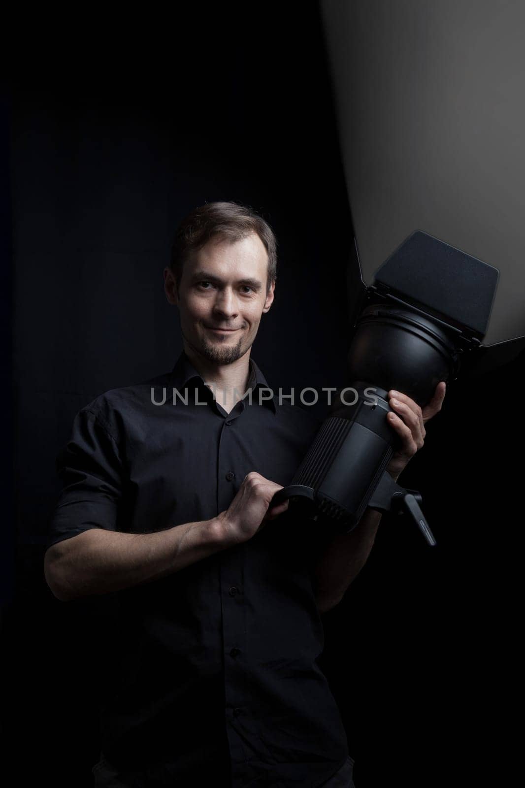 Portrait of a gaffer man with a professional lighting head device in his hands on a black background.