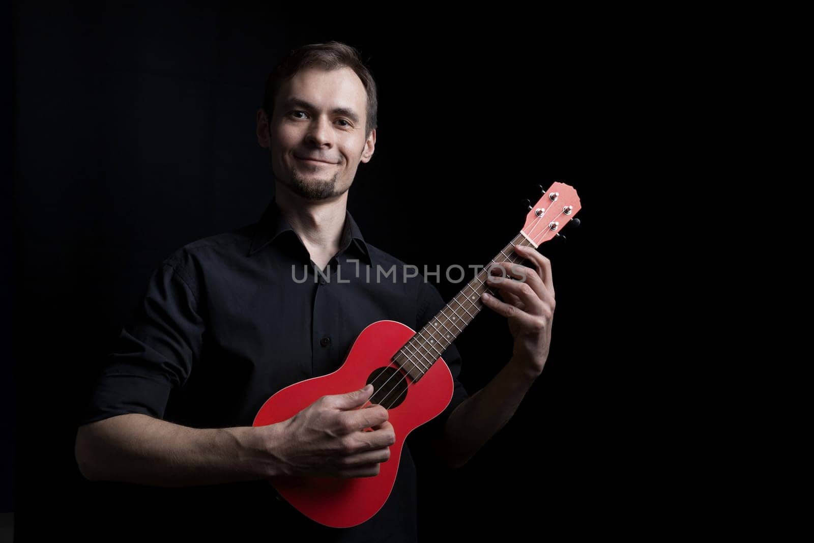 Handsome smiling low key man portrait playing on fun red ukulele guitar over black background.