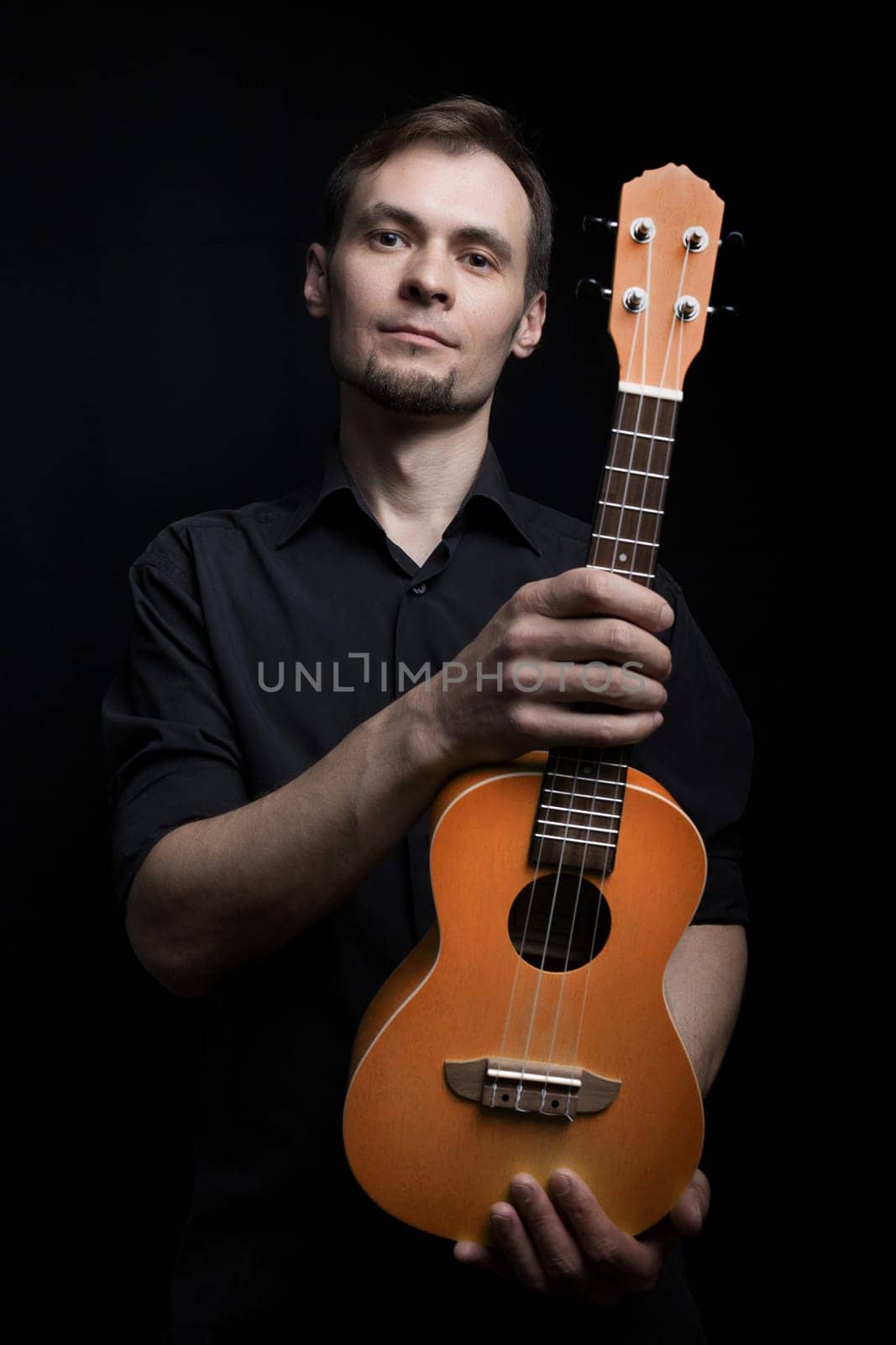 Handsome young low key man portrait with ukulele guitar over black background.