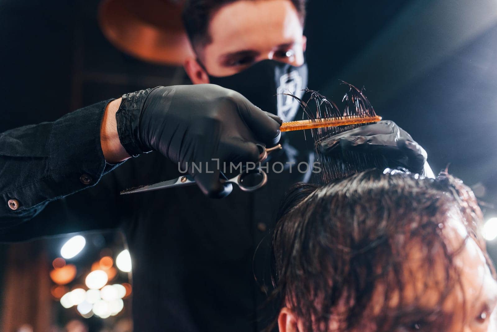 Young bearded man sitting and getting haircut in barber shop by guy in black protective mask.