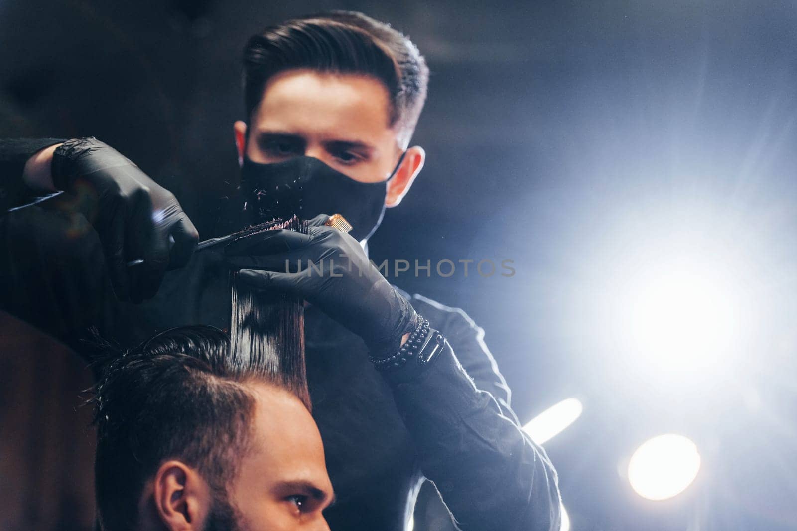 Young bearded man sitting and getting haircut in barber shop by guy in black protective mask.