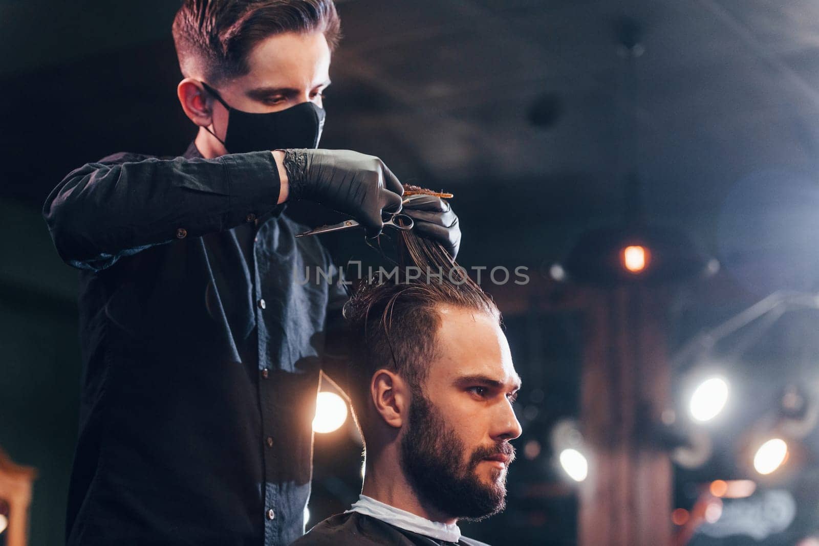 Young bearded man sitting and getting haircut in barber shop by guy in black protective mask.