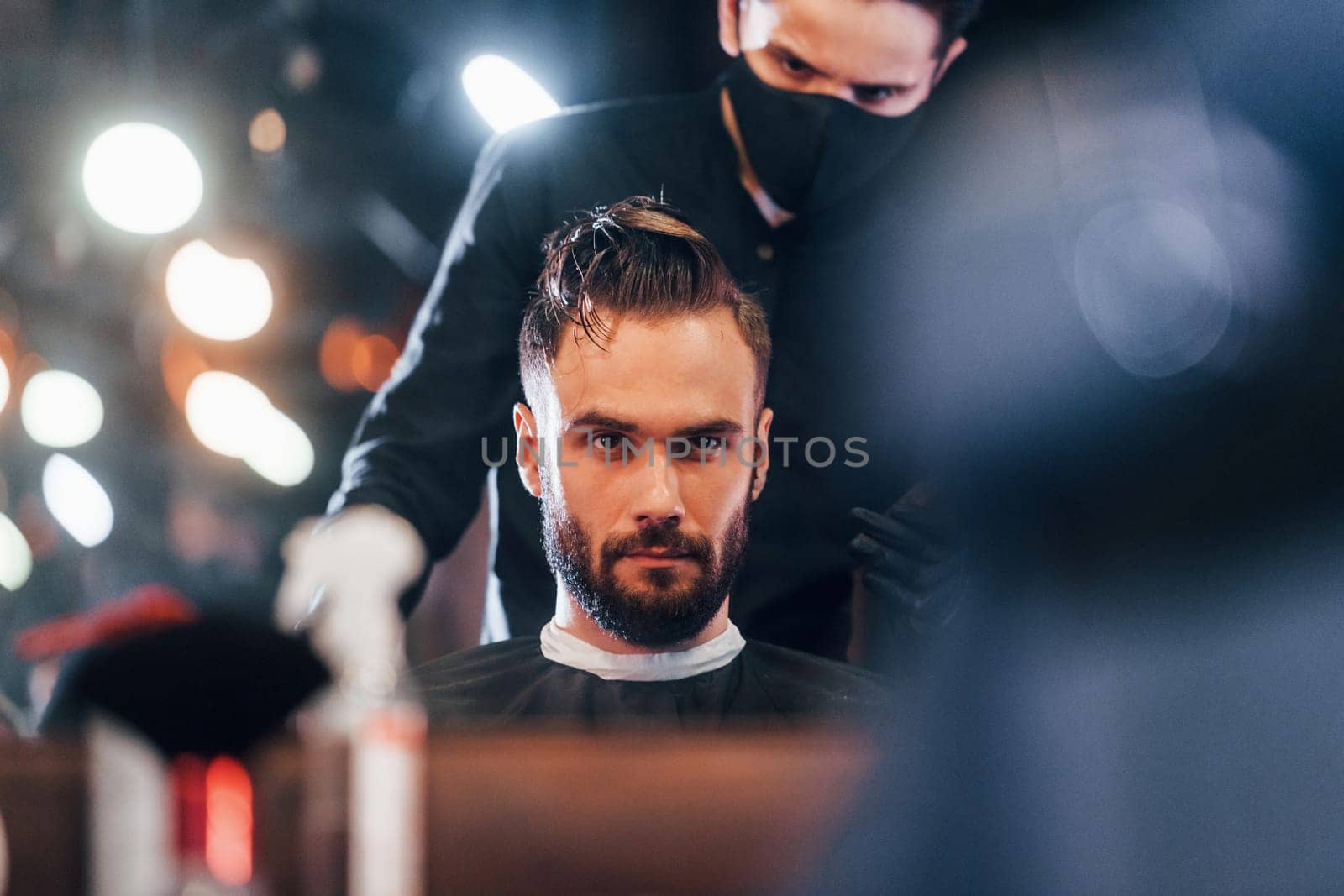 Front view of young bearded man that sitting and getting haircut in barber shop by guy in black protective mask.