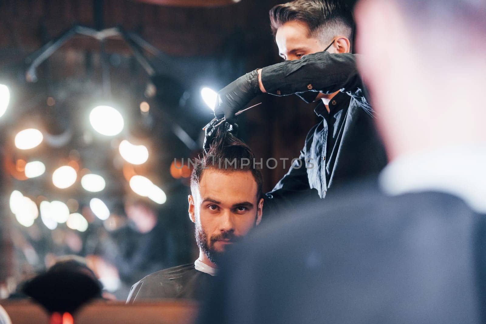 Front view of young bearded man that sitting and getting haircut in barber shop by guy in black protective mask.