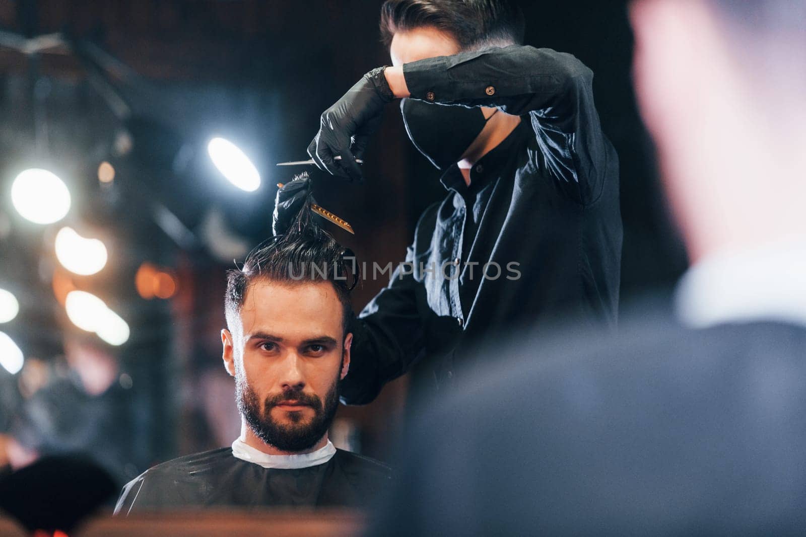Front view of young bearded man that sitting and getting haircut in barber shop by guy in black protective mask by Standret