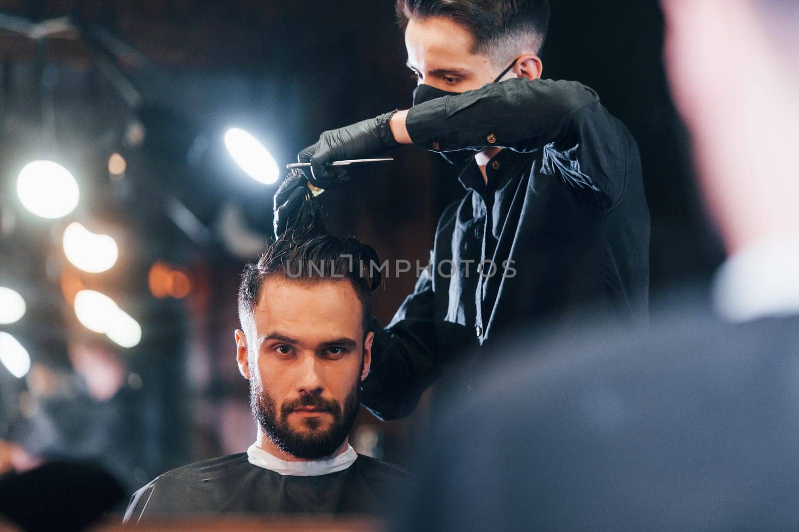 Front view of young bearded man that sitting and getting haircut in barber shop by guy in black protective mask.