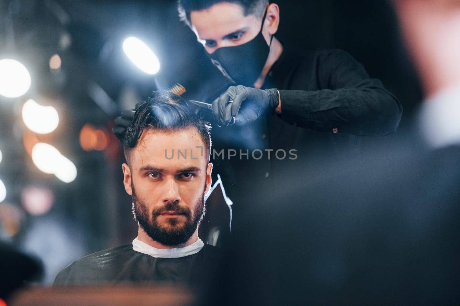 Front view of young bearded man that sitting and getting haircut in barber shop by guy in black protective mask by Standret