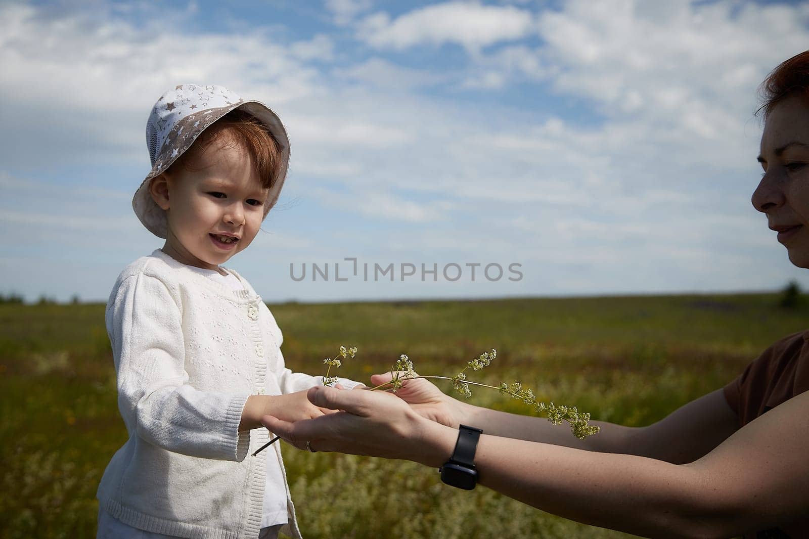 Happy female family with mother and daughter on green and yellow meadow full of grass and flower. Woman with red hair and blonde girl having fun, joy and hugs in sunny summer day. Concept family love by keleny