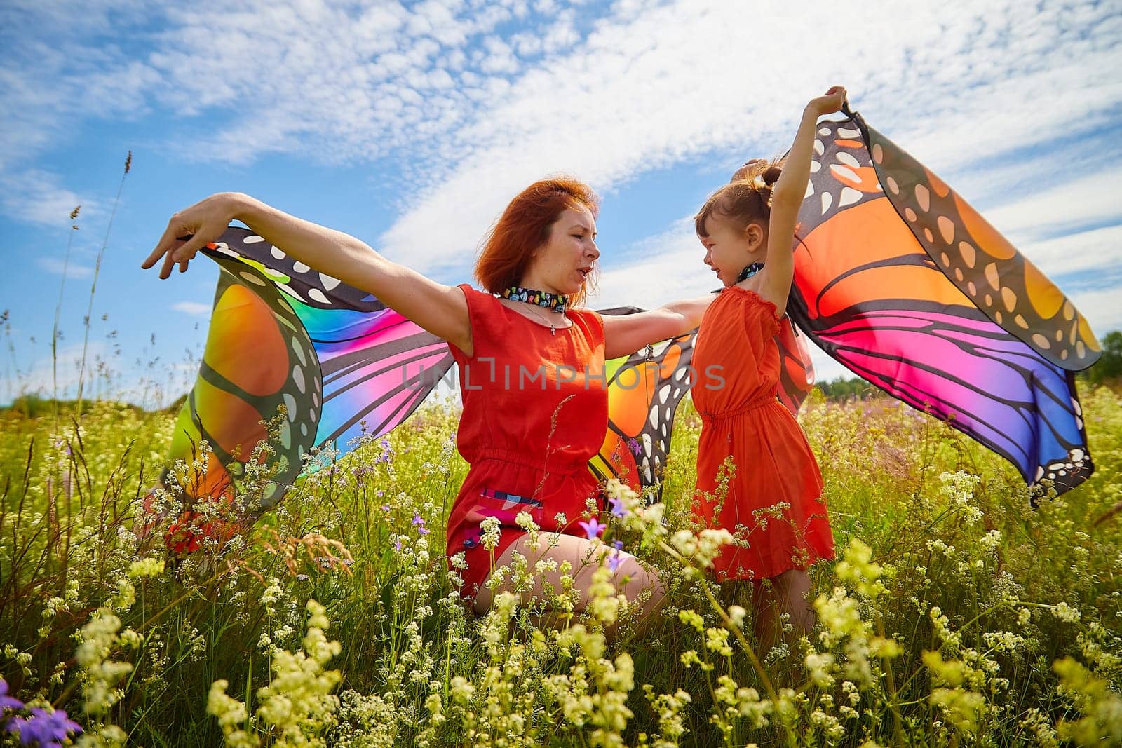 Happy female family with red haired mother and daughter with bright butterfly wings having fun on green and yellow meadow full of grass and flower in sunny summer day. Concept family love by keleny