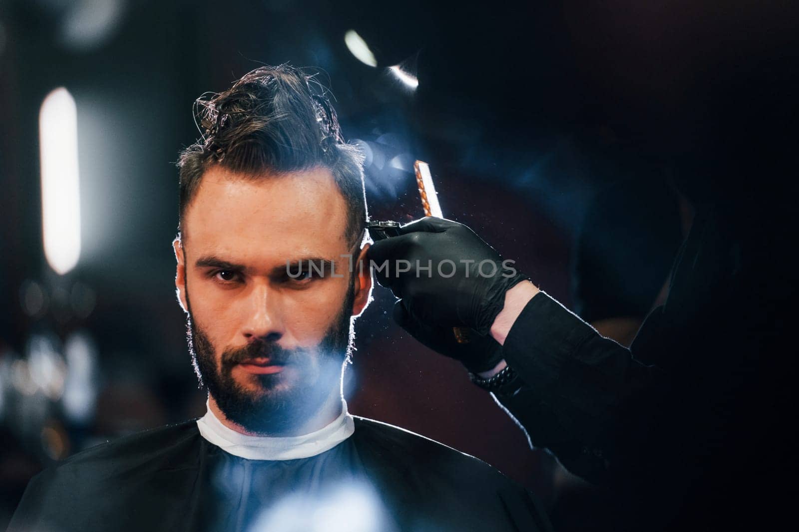 Young bearded man sitting and getting haircut in barber shop.