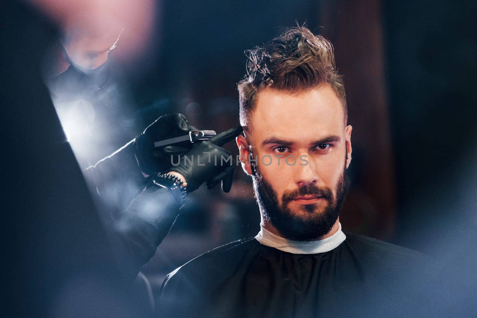 Young bearded man sitting and getting haircut in barber shop by Standret